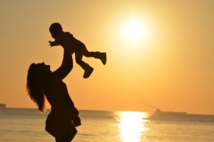 woman holding her baby in the air as the sun sets over a body of water behind her
