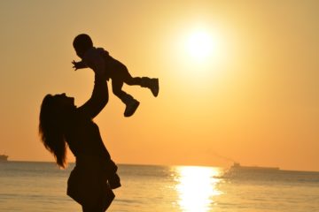 woman holding her baby in the air as the sun sets over a body of water behind her
